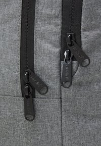 Šedý látkový batoh s černými zipy a táhly. Materiál má texturovaný vzhled a minimální detaily šití. Jsou viditelné tři zipové otvory.