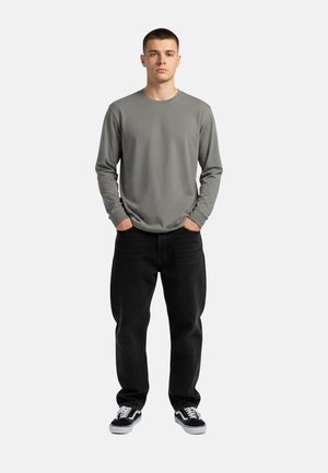 Junger Mann steht mit den Händen in den Taschen, trägt ein graues Langarmshirt, schwarze Jeans und schwarz-weiße Turnschuhe vor weißem Hintergrund.