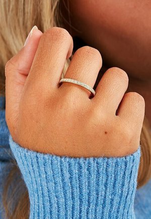 Goldring mit kleinen klaren Steinen, runde Form, getragen an einer Hand mit heller Haut; Hand teilweise sichtbar neben einem blauen gerippten Pullover.
