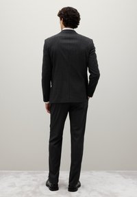Traje de rayas gris oscuro con un diseño ajustado, que cuenta con una única abertura y una tela suave. Los pantalones tienen un corte a medida. Unos zapatos negros completan el look.