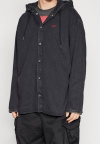 Person som bär en svart huvad jeansjacka med tryckknappar och svarta cargo-byxor, händer delvis synliga på sidorna.