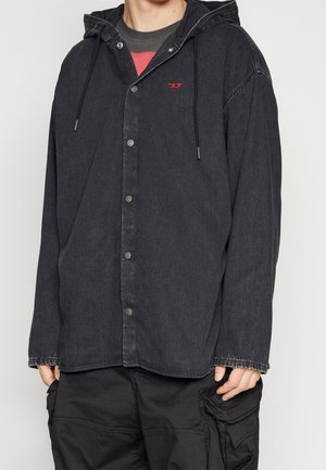 Persona che indossa un giubbotto di denim nero con cappuccio e bottoni a pressione, abbinato a pantaloni cargo neri, con le mani parzialmente visibili ai lati.