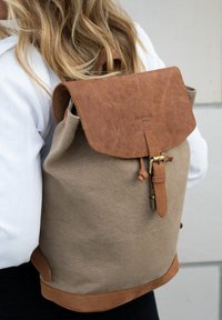 Person wearing beige canvas backpack with brown leather flap, belt buckle, and embossed "Holzrichter Berlin" logo, against a light wall background.