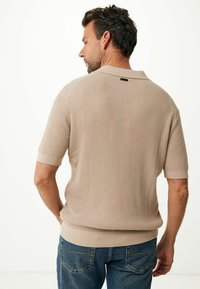 Polo de punto beige de manga corta con un patrón texturizado, cuello y dobladillo acanalados, combinado con jeans azules; vista trasera visible.