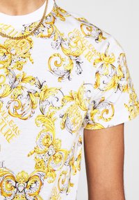 Hvid t-shirt med detaljeret gul og sort barokmønster med tekstaccents, korte ærmer og rund halsudskæring.