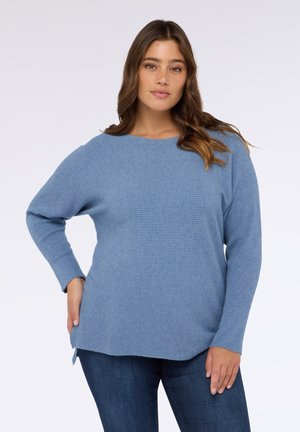 Maglione azzurro chiaro a coste con un'ampia scollatura e maniche lunghe, dotato di spacchi laterali all'orlo per facilitare i movimenti. Abbinato a jeans scuri.