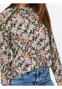 Blusa floreale con colletto alto, maniche lunghe e vivaci motivi di fiori lilla, gialli e bianchi su uno sfondo verde.