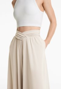 Beige weitbeinige Hosen mit einem gerafften Bund und seitlichen Taschen, aus weichem Stoff gefertigt, kombiniert mit einem weißen gerippten Cropped Top.