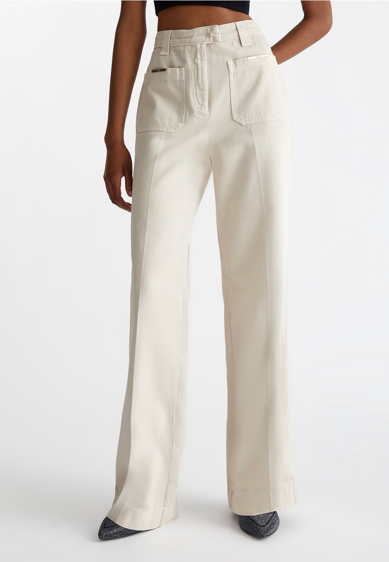 Pantalon beige taille haute à jambes larges avec poches plaquées sur le devant, porté avec des chaussures noires pointues et un top court noir.
