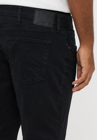 Μαύρο τζιν από denim με slim γραμμή, πλάγια πίσω τσέπη σε γωνία, δερμάτινη ετικέτα στη μέση και διακριτικές ραφές.