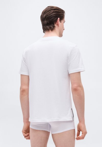 Άνδρας με απλό λευκό μπλουζάκι και λευκά εσώρουχα μποξέρ, όρθιος, γυρισμένος πλάτη, μπροστά σε απλό ανοιχτό φόντο.