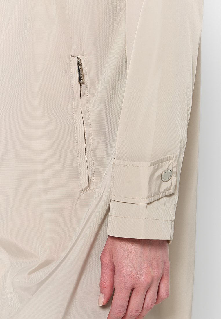 Veste légère beige avec une poche zippée et un poignet boutonné. Le tissu a une texture subtile, mettant en avant un design minimaliste.