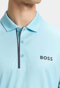 Jasnoniebieska koszulka polo z teksturowanym wzorem, kontrastowym ciemnym zamkiem błyskawicznym, dwoma guzikami oraz czarnym logo "BOSS" po lewej stronie.