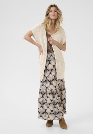Beige Strickjacke mit kurzen Ärmeln, die über einem braunen Oberteil und einem gemusterten Maxirock mit Palmenblatt-Designs in Schwarz und Beige getragen wird.