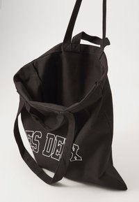 Black canvas tote bag with two handles, featuring white text on one side. The bag has a spacious, open interior and a tapered bottom.