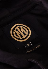 Zwarte hoodie-interieur met een opvallend gouden geborduurd rond logo, gemarkeerd met maat 'S' en een 'Made in Europe' label. Zachte textuur, minimalistisch ontwerp.
