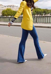 Žlutý blazer přes pruhovanou košili s dlouhým rukávem, rozšířené modré denimové džíny a bílé boty. Venku, na chodníku s stromy v pozadí.