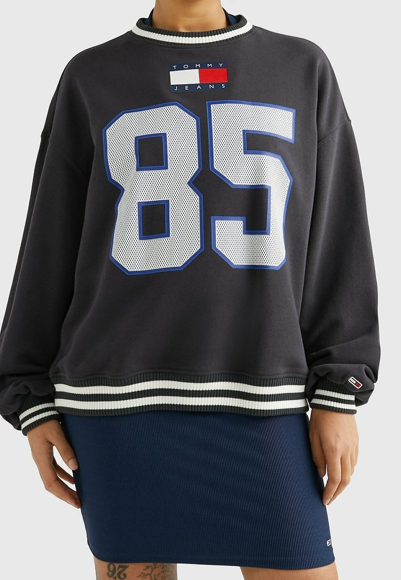 Personne portant un sweat-shirt Tommy Jeans gris foncé avec le numéro 85 en maille blanche et des poignets rayés, associée à une jupe côtelée bleu marine.
