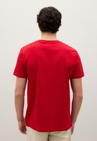 Uomo con capelli ricci scuri che indossa una semplice maglietta rossa a maniche corte e pantaloni beige, in piedi di fronte a una parete bianca.