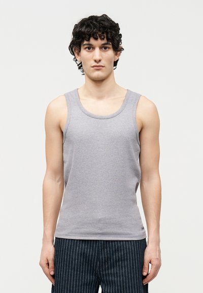 Νεαρό άτομο με κοντά σγουρά μαλλιά που φορά γκρι αμάνικο τοπ και σκούρο παντελόνι με λεπτές ρίγες, στέκεται μπροστά σε απλό φωτεινό φόντο.