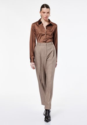 Donna con indosso una camicia di satin marrone con bottoni, pantaloni beige a vita alta su misura, stivaletti neri e orecchini a cerchio dorati su sfondo bianco.
