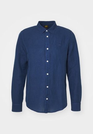 Granatowa koszula z długim rękawem, zapinana na guziki, z białymi guzikami i subtelnym haftowanym logo na piersi. Miękka textura, prosta sylwetka.