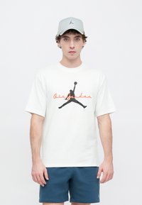 Λευκό βαμβακερό μπλουζάκι με μαύρο λογότυπο Air Jordan και κόκκινο κείμενο, σετ με μπλε σορτς και γκρι καπέλο με μαύρο λογότυπο.