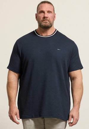 Homme barbu portant un t-shirt bleu marine à manches courtes et un pantalon beige, debout devant un fond clair uni.