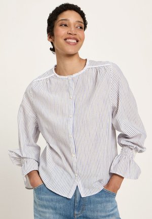 Smiling person with short curly hair wearing a white blouse with thin blue stripes and puffed sleeves, hands in pockets of blue jeans.