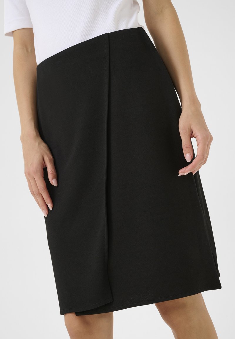 Femme portant une jupe portefeuille noire longueur genou avec un haut blanc à manches courtes, debout avec les mains relâchées le long du corps.