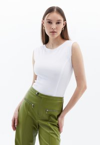 Bílý bezrukávový žebrovaný top spárovaný s vysokými zelenými kalhotami s kapsami na zip, hladkou texturou a přiléhavým střihem.
