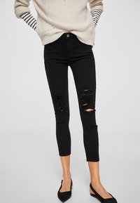 Vaqueros skinny negros con desgarros desgastados en las rodillas, con una silueta ajustada y una tela suave y texturizada. Combinados con un suéter ligero.