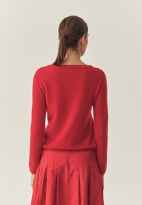 Sarkans džemperis ar mīkstu, struktūru apdari, ar garām piedurknēm un apaļu kakla izgriezumu, saskaņots ar atbilstošu sarkanu plisētu svārku.