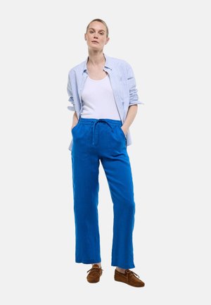 Femme debout les mains dans les poches, portant un débardeur blanc, une chemise rayée bleue, un pantalon bleu vif et des mocassins marron.