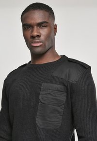 Suéter negro de punto con hilo texturizado, acentos de hombros acolchados y un bolsillo delantero con solapa. Sutil contraste entre materiales.