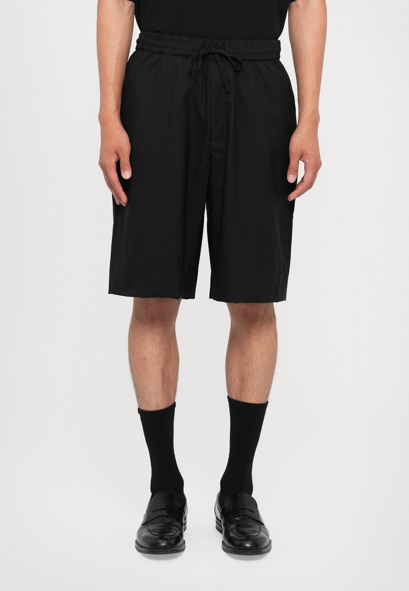 Shorts noirs en matériau léger avec une taille élastique et un cordon de serrage ajustable. Portés avec des chaussettes et des chaussures noires.