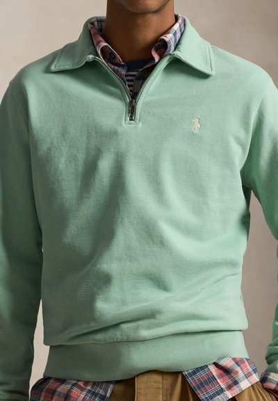 Άνδρας που φοράει φούτερ μισό φερμουάρ σε μέντα πράσινο χρώμα πάνω από πουκάμισο κόκκινο και μπλε καρό, με καφέ παντελόνι, μπροστά σε ουδέτερο φόντο.