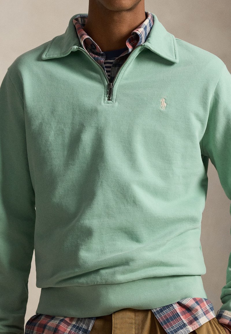 Man wearing mint green quarter-zip pullover over red and blue plaid shirt, with brown pants, against a neutral background.