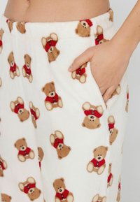 Fleecové pyžamové kalhoty s bílým základem a opakujícím se vzorem medvídků v béžové a červené barvě. Obsahují boční kapsy. Měkká textura.