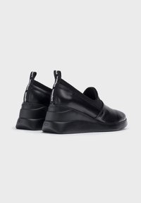 Zapatos de piel negra sin cordones con una parte superior suave, punta redondeada y suela de cuña de goma. Presentan lengüetas con marca en los talones.