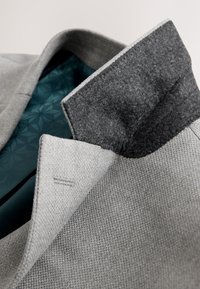 Col de manteau gris texturé avec doublure intérieure gris foncé rabattue, révélant un tissu à motifs bleu sarcelle à l'intérieur du manteau.