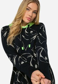 Haut à capuche noir avec un motif floral, doublure de capuche verte et cordons. Il présente des manches longues, des poignets côtelés et une coupe décontractée.