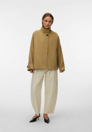 Ženska stoji, oblečena v bež jakno z visokim ovratnikom, široke krem barve hlače in črne salonarje z vbočenimi konicami pete, na belem ozadju.