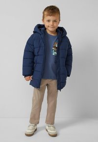 Granatowa pikowana kurtka puchowa z kapturem, noszona na niebieskiej koszuli z nadrukiem graficznym, w połączeniu z beżowymi spodniami i białymi trampkami.