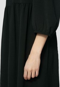 Schwarzes Kleid mit geraffter Taille, weicher Textur und puffartigen Ärmeln. Nahaufnahme einer Hand, die auf dem Stoff ruht, um die Details hervorzuheben.