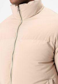 Lichtbeige pufferjas met een hoge kraag en ritssluiting, voorzien van een gladde, matte textuur en zijzakken.