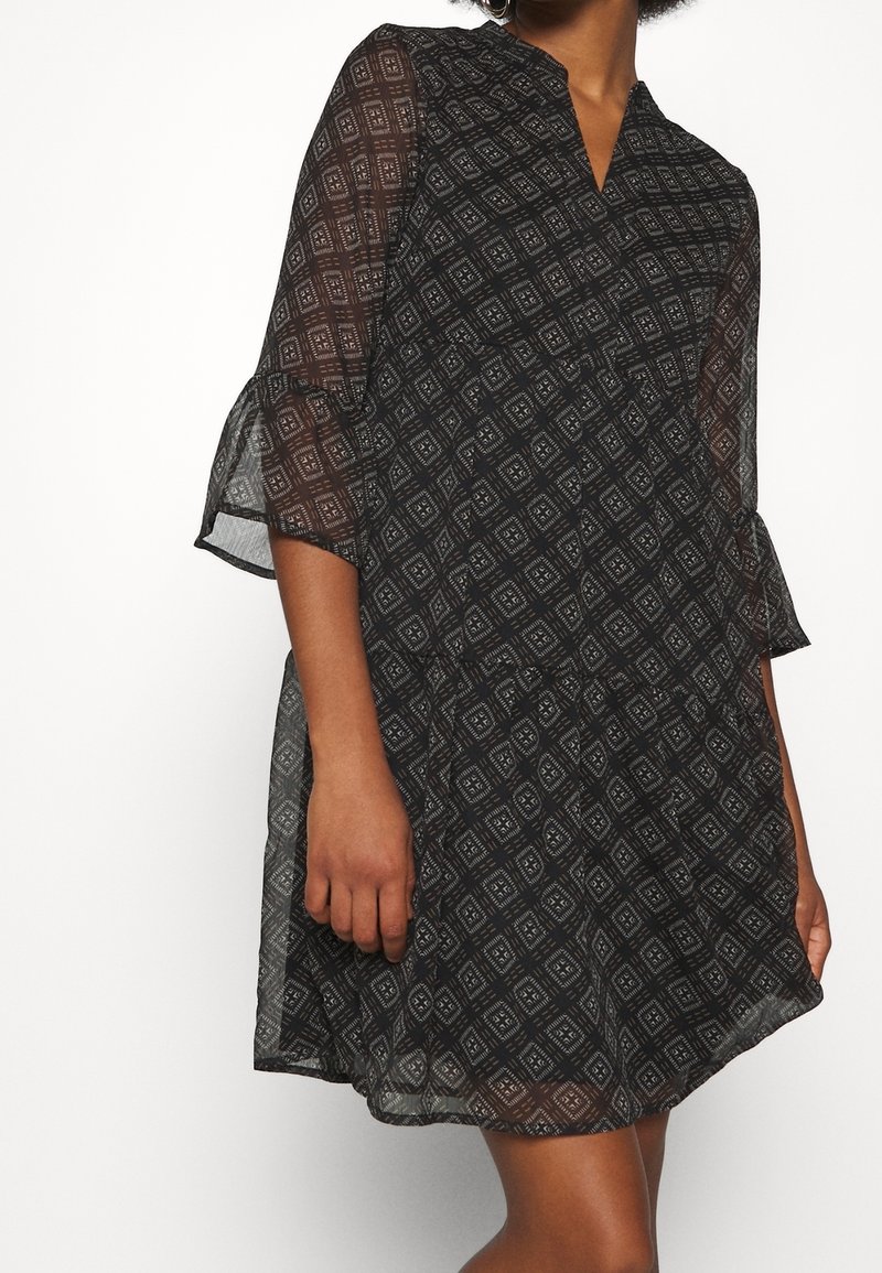 Femme portant une robe noire semi-transparente avec des motifs géométriques blancs en forme de losange et des manches cloche trois-quarts, debout sur un fond blanc.