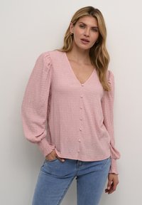 Blusa rosa, testurizzata, con scollo a V, maniche lunghe a sbuffo e bottoni frontali. Accentata con polsini arricciati. Abbinata a jeans azzurri chiari.