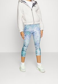 Chaqueta de forro polar con cremallera en color crema, con un interior estampado, combinada con leggings capri azul tie-dye. El modelo lleva zapatillas blancas y calcetines.