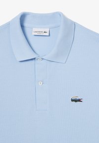 Ljusblå pikétröja i texturerad tyg, med en tre knappars knappslå och en liten broderad krokodil-logotyp på bröstet.